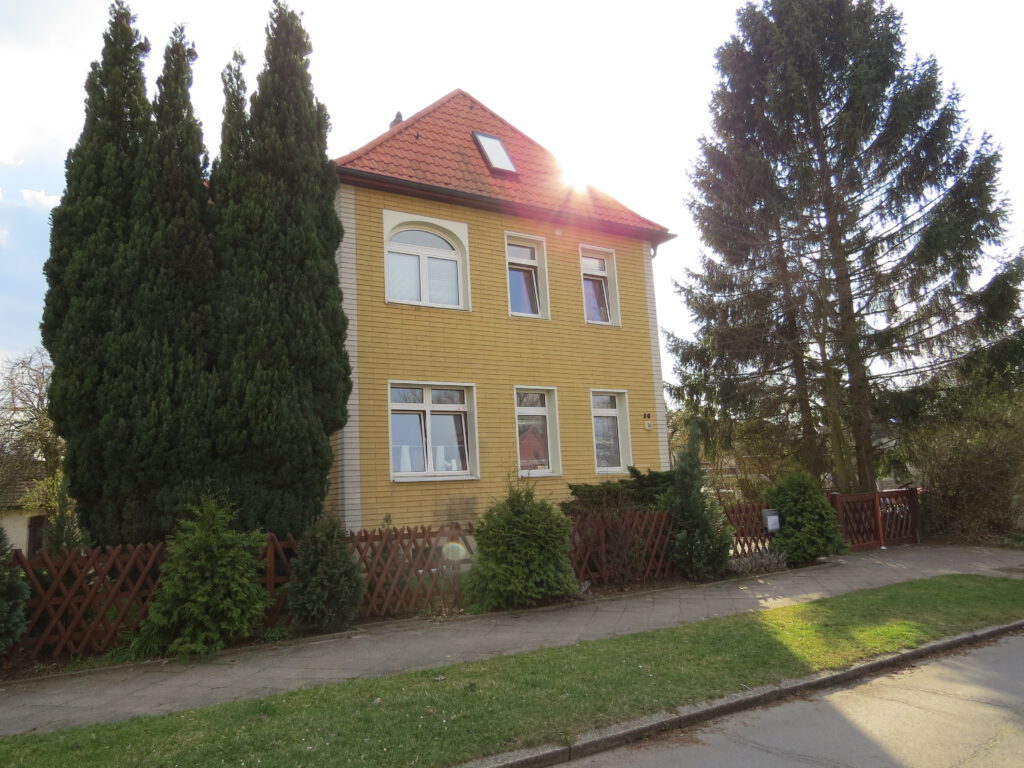 Villa MITTENDRIN - Betreutes Jugendwohnen - Zweistöckiges Haus mit Spitzdach - gelbe Fassade, rotes Dach - rechts und links vom Haus stehen Bäume, über dem Dach blitzt die Sonne ins Objektiv