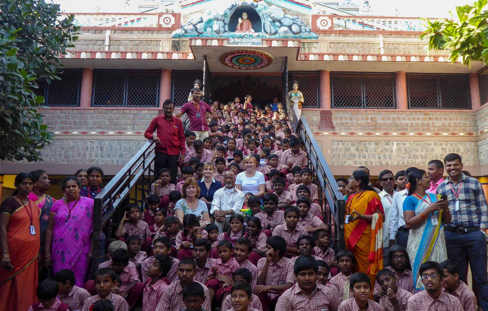 Indien. Viele Menschen sitzen auf einer Treppe vor einem Indischen Gebäude. Alle tragen Schuluniform. In der Mitte der Gruppe sitzen die Geschäftsführerinnen von MITTENDRIN leben e.V. - Internationale Hilfe