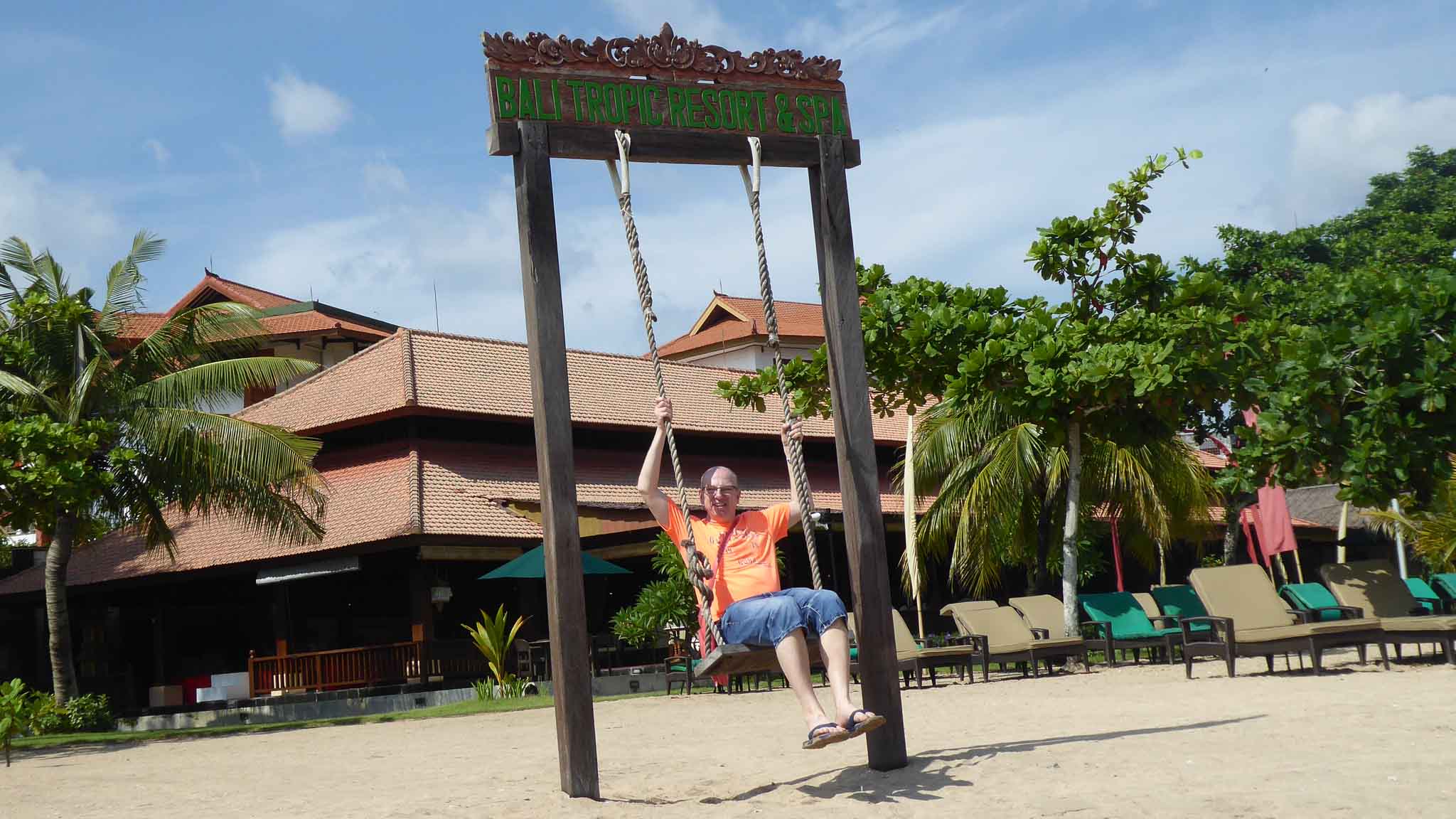 Klientenreise Bali - ein Mann auf einer großen Schaukel am Strand. Im Hintergrund ein Hotel, welches als Tempelanlage gestaltet ist