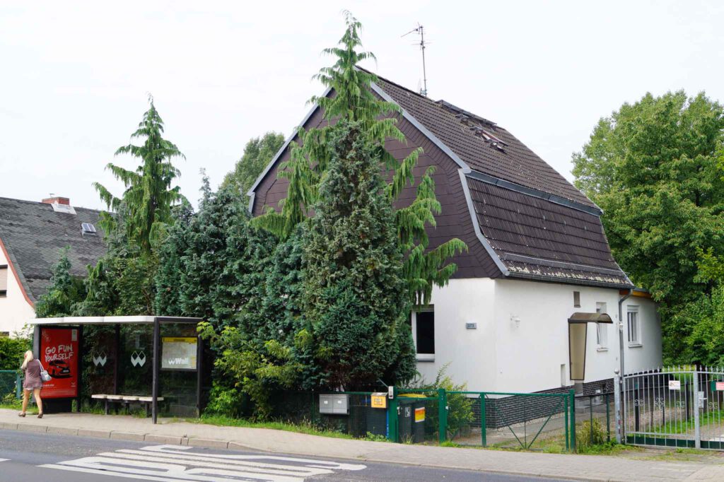 Intensiv betreutes Einzelwohnen Hönower Straße. Einfamilienhaus mit weißer Fassade und dunklem Dach mit Garten. Vor dem Haus stehen Nadelbäume