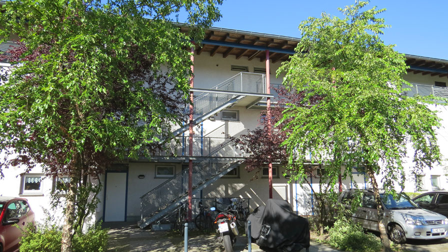 TWG Waldowpark von außen. Wohnhaus mit flachem Dach und hellblauer Fassade. Davor zwei Bäume und parkende Autos