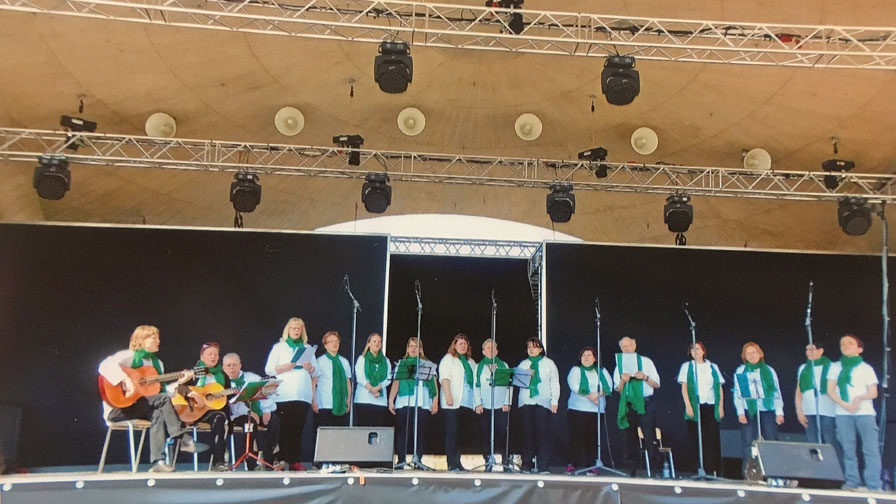 Singegruppe: 16 Personen stehen nebeneinander auf einer großen Bühne und singen. Alle tragen schwarze Hosen, weiße Hemden und grüne Halstücher