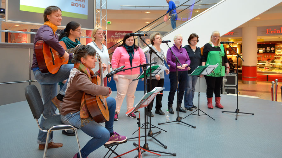Singegruppe: Im Einkaufszentrum Eastgate singt die Singegruppe auf einer Veranstaltungsfläche. Es sind 6 Sängerinnen und zwei Gitarrenspielerinnen zu sehen. Im Hintergrund Geschäfte und eine Rolltreppe