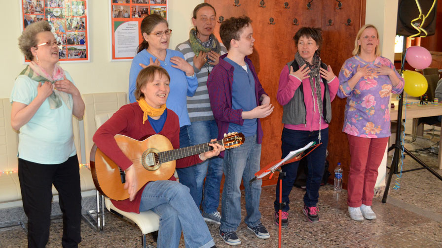 Singegruppe: 6 Sängerinnen und Sänger stehen vor einer Holzwand und singen. Zusätztlich sitzt eine Gitarrenspielerin auf einem Stuhl