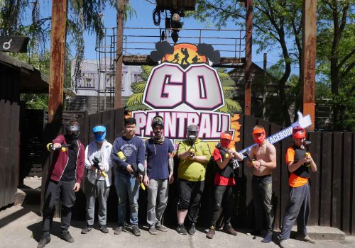 8 Personen beim Paintball. Alle stehen mit Maske und ihren Markierern vor einem großen Schild auf den "Go Paintball" steht