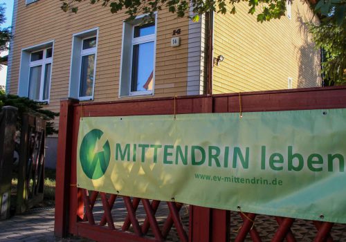 Foto von der Villa. Am roten Gartentor hängt ein Banner von MITTENDRIN leben e.V. im Hintergrund das Haus mit der gelben Fassade