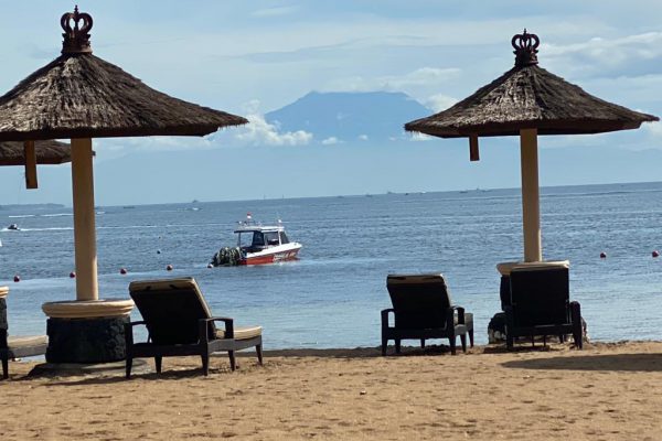 Betreute Reise Bali. Strand mit 2 Schirmen aus Stroh und Sonnenliegen. Im Hintergrund fährt ein Boot.