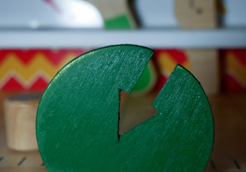 Einzelfallhilfe: Eine Werkbank für Kinder. Im Schraubstock im Vordergrund ist ein grüner Holzkreis mit einem eingesäckten Pfeil befestigt. Im Hintergrund sieht man verschwommen eine Holzsäge und einen Holzmaulschlüssel, die jeweils einen lächelnden Smiley aufgedruckt haben.