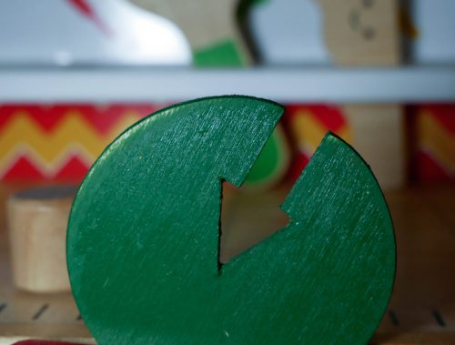 Werkbank Einzelfallhilfe: Eine Werkbank für Kinder. Im Schraubstock im Vordergrund ist ein grüner Holzkreis mit einem eingesäckten Pfeil befestigt. Im Hintergrund sieht man verschwommen eine Holzsäge und einen Holzmaulschlüssel, die jeweils einen lächelnden Smiley aufgedruckt haben.